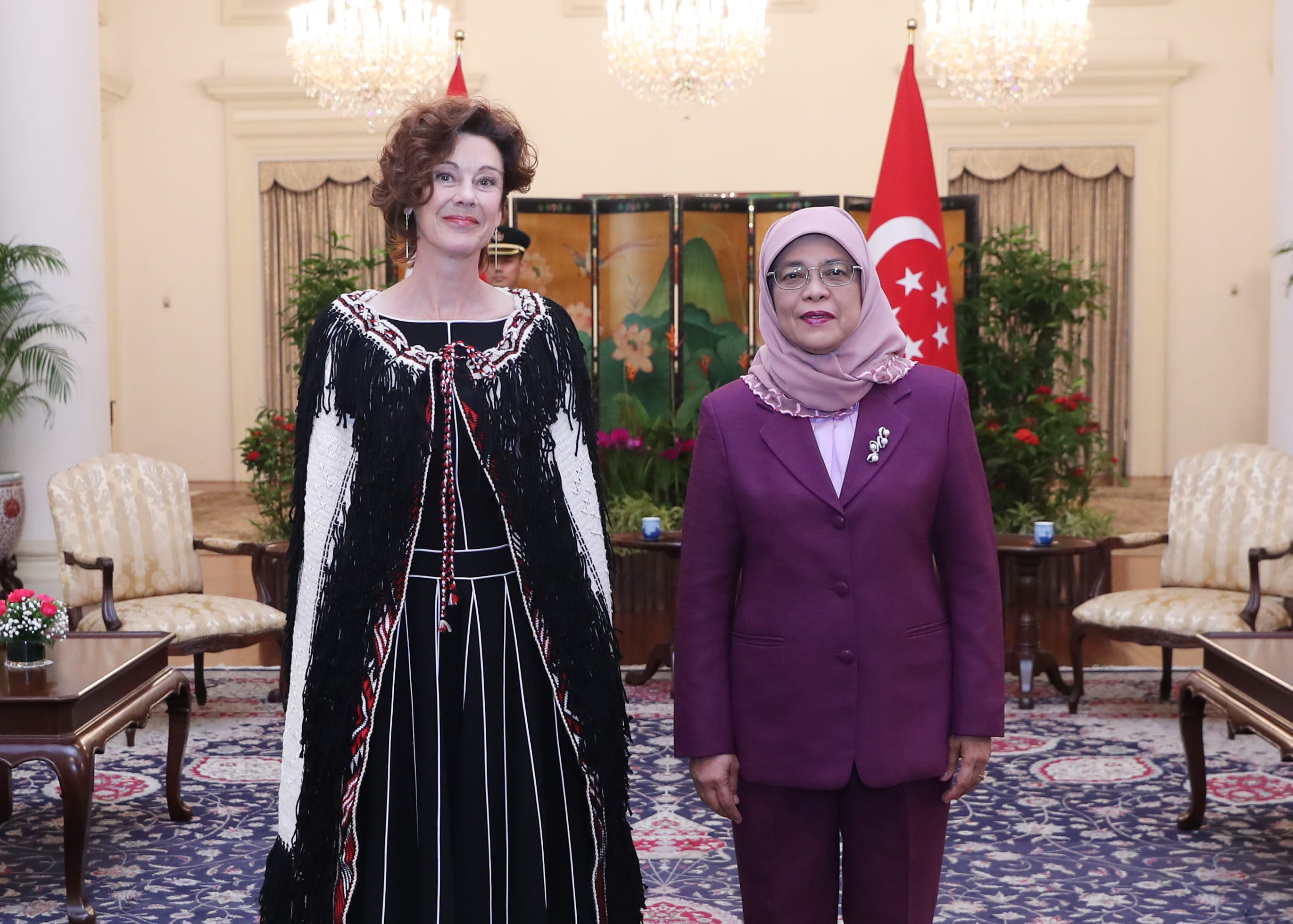 Two women standing indoors, Singapore flag in background.
